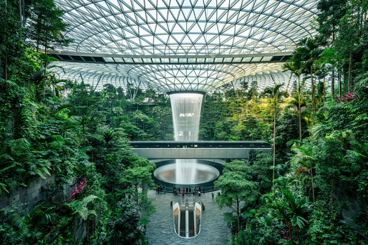 Architectural interior with glass ceiling, lush vegetation, and waterfall cascading over circular platform.