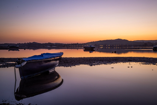 Blue and dark fishing boat anchored in still water at dawn, with distant shoreline and bridges visible under purple and orange sky.
