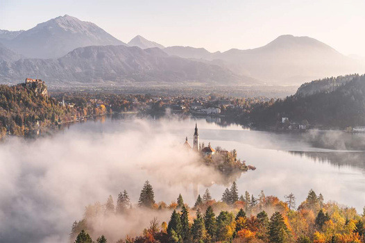 Church on a forested island surrounded by autumn landscape and fog-covered lake with mountains beyond.