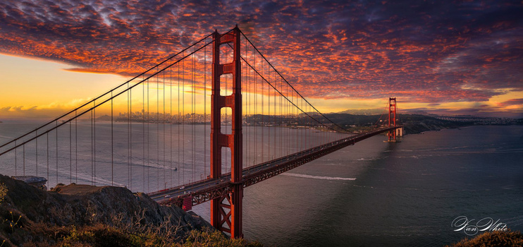Golden Gate Bridge silhouetted against dramatic orange and purple sunset sky with calm bay waters below.