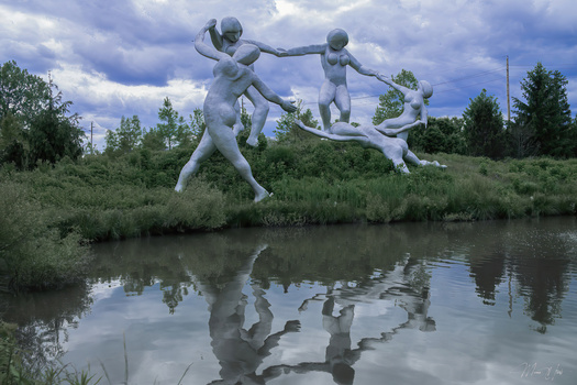 Large white sculptural figures suspended above a reflective river with forested banks and cloudy sky.