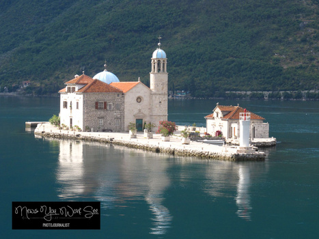 Orthodox church with bell tower on small rocky island in turquoise water surrounded by forested mountains.