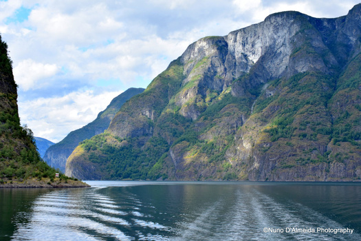 Steep rocky mountain peaks frame a calm alpine lake with forested slopes and a small boat wake visible on the water.