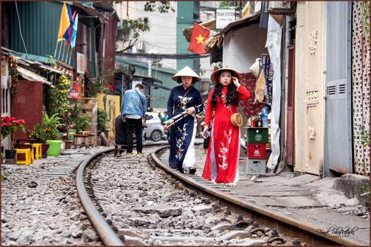 Woman in red áo dài walks along narrow street with railroad tracks running through urban neighborhood.