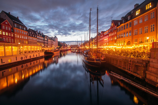 Illuminated waterfront canal at dusk with moored sailboats and colorful historic buildings reflected in still water.