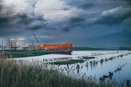 Orange industrial barge moored in shallow water with wooden pilings and industrial facility in background under dramatic storm clouds.