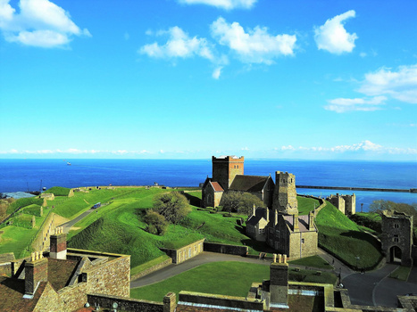 Medieval hilltop castle with stone towers overlooking blue ocean and green grassland under clear sky.
