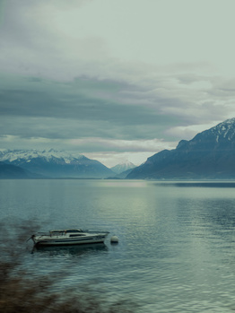 Small wooden boat anchored in calm alpine lake with snow-capped mountains under overcast sky.