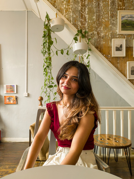 Woman with long dark hair wearing a red sleeveless top and white pants, seated at a table in a bright interior space.