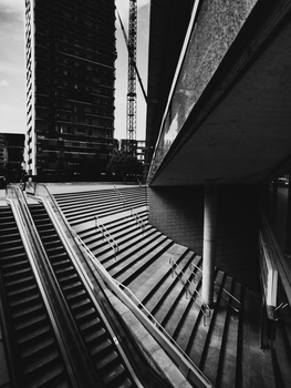 Urban architectural perspective with diagonal lines of building facades and stairs converging toward a distant skyscraper in black and white.
