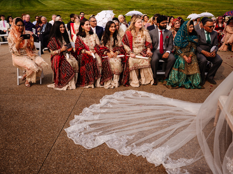 Wedding guests in traditional South Asian attire seated for ceremony, with bride's ornate veil train visible in foreground.