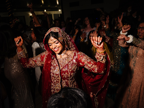 Bride in ornate red and gold embroidered lehenga dancing at wedding celebration surrounded by guests.