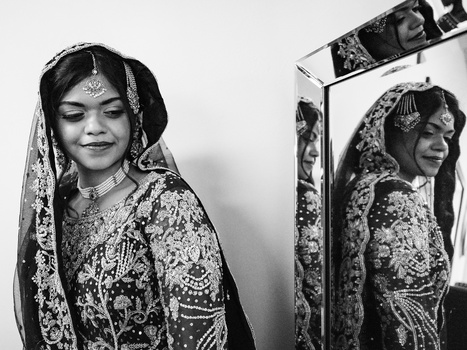 Woman in ornate traditional wedding attire with intricate embroidery and veil, photographed in black and white beside a mirror reflection.