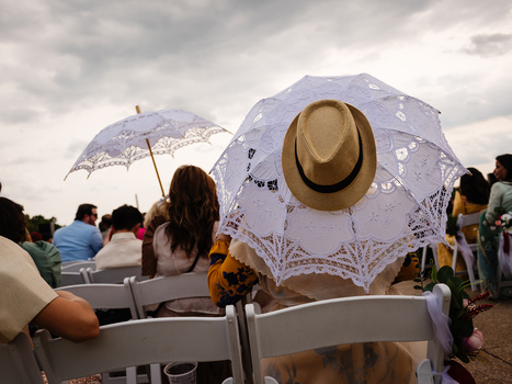 Woman holding ornate lace parasol and tan hat at outdoor event, seated among crowd on white chairs.