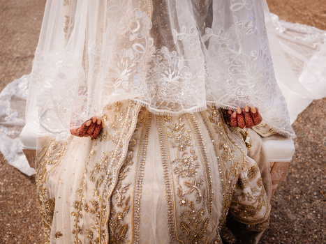 Bride wearing ornate gold-embroidered lehenga with delicate henna on her hands, sheer dupatta draped overhead.