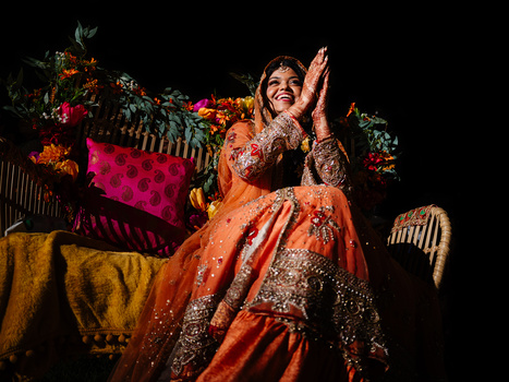Woman in ornate orange and gold Indian bridal attire clapping with joy against a dark backdrop with vibrant florals and fabrics.