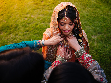 Bride in ornate traditional wedding attire smiling while being adorned with jewelry by family members.