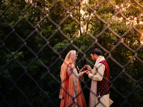 Two people in traditional South Asian wedding attire exchanging rings at a chain-link fence with golden-hour sunlight and green foliage behind.