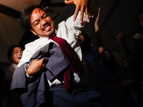 Man in white shirt and burgundy tie gestures expressively while speaking, surrounded by audience members in dim lighting.