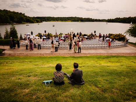 Two people seated on grass overlooking a waterside wedding ceremony setup with guests and white chairs.