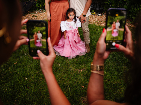 Young girl in pink skirt and white top standing on grass while multiple people photograph her with smartphones.