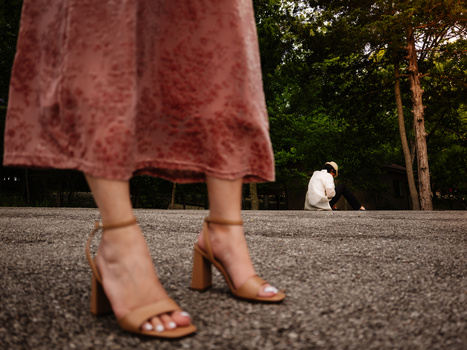 Woman's legs in tan heeled sandals and mauve dress standing on pavement, with blurred figure and trees in background.