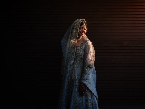 Woman in ornate beaded gown and lace veil standing against dark background with side lighting.
