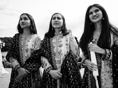 Three women in ornate embroidered tunics stand together looking upward in black and white.