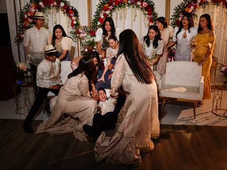 Wedding guests in formal attire gather around a seated couple during an indoor ceremony with floral garland decorations.