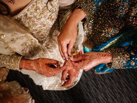Hands with henna mehndi designs applying paste to another person's arm during a traditional application ceremony.
