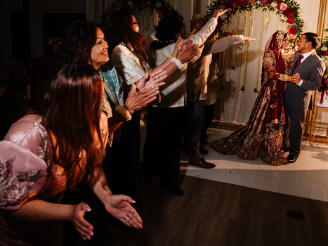 Wedding guests in formal attire celebrate as a couple dances in an elegant indoor venue decorated with garland.