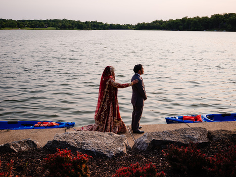 Couple in formal attire standing on rocks by a waterside at golden hour, with blue boats moored nearby.