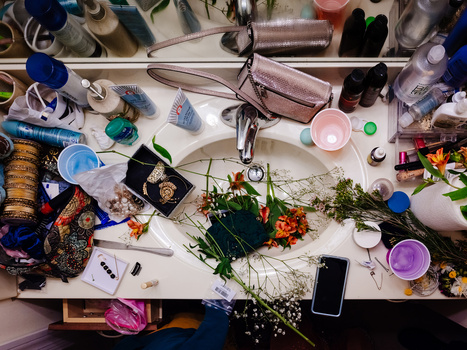 Overhead view of a creative workspace with botanical specimens, cosmetic products, and artistic materials arranged on a white surface.