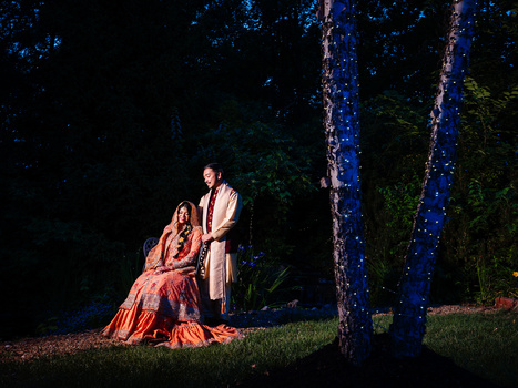 Two figures in orange and tan robes sit beneath illuminated blue trees at night.