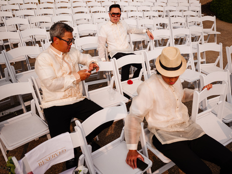 Three men in white shirts seated among white folding chairs at an outdoor event.