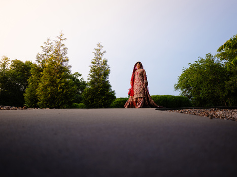 Woman in red standing alone on dark asphalt road with green trees and clear sky behind her.