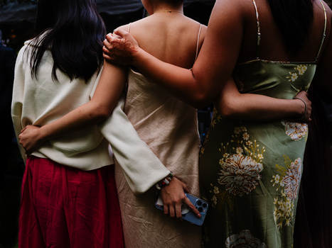 Three women with arms around each other from behind, wearing casual and floral clothing against a dark background.