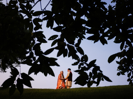 Two figures in orange robes standing on a grassy horizon, framed by dark silhouetted foliage.