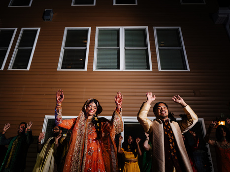 Wedding guests in traditional attire dancing with arms raised against a wooden building facade at night.