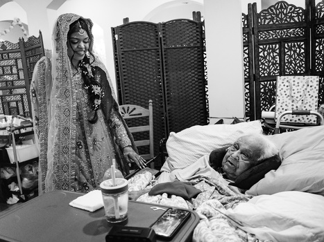 Woman in ornate traditional dress and veil standing beside a person resting on a bed in a black and white photograph.