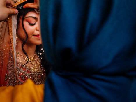 Woman in ornate traditional dress with gold embroidery holding sheer red fabric against blue background.