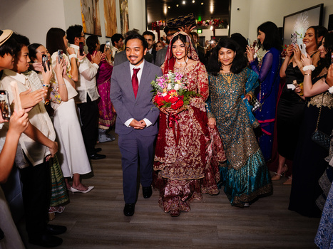 Wedding procession with groom in navy suit and bride in red embroidered lehenga holding flowers, flanked by guests in formal attire.