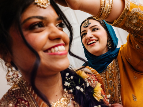 Two women wearing traditional South Asian wedding attire with gold embroidery, jewelry, and bindis, smiling at the camera.