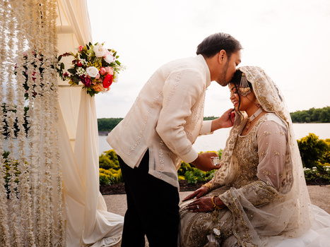 Groom in cream jacket kissing bride's hand during outdoor wedding ceremony by water.