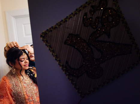 Woman in embellished orange garment poses beside dark textured wall art with beaded details.