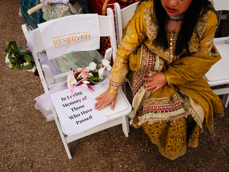 Woman in gold patterned dress crouches beside white reserved chair with flowers and handwritten sign.
