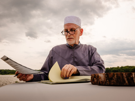 Older man with glasses examining a fish against a cloudy sky backdrop.