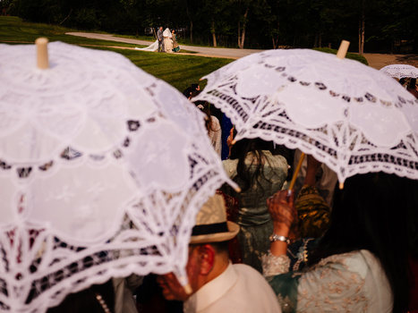 People holding white lace umbrellas at an outdoor evening event with green lawn and trees in background.