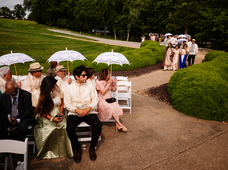 Outdoor wedding ceremony with guests holding white umbrellas seated on a tree-lined lawn.