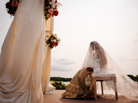 Bride in white veil kneeling at wooden bench during outdoor sunset ceremony with floral arch.